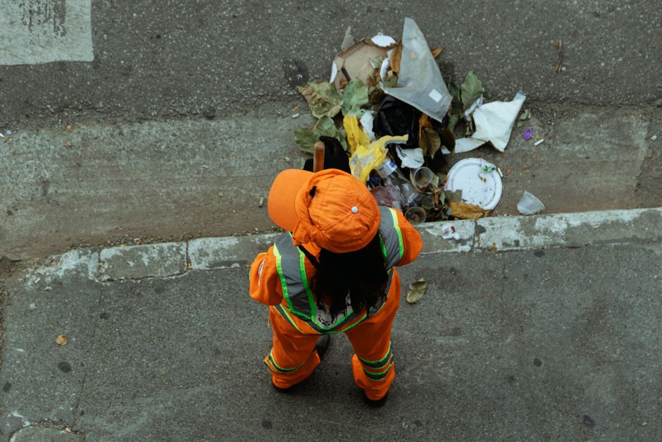 A close-up view of multiple large white plastic trash bags filled with various household items, sealed with orange tie wraps, and placed on a paved outdoor surface. The bags contain soft furnishings,