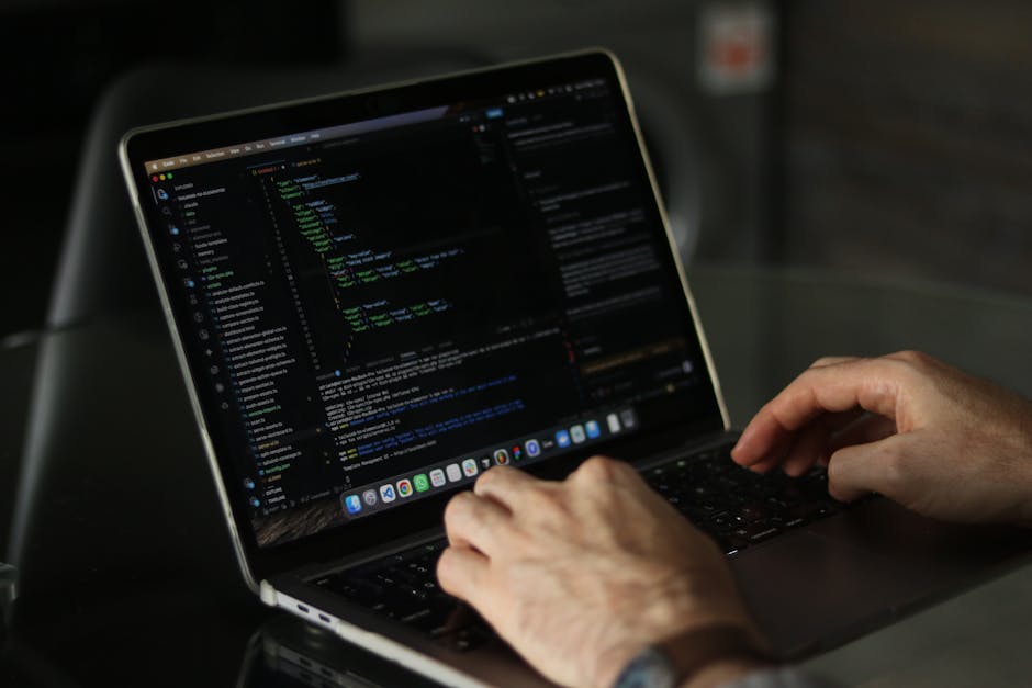 A person using a laptop on a wooden surface, with their right hand resting on the keyboard and wearing a wristwatch. The laptop screen displays lines of code in a text editor. This image illustrates t